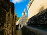 Ancient stone structures and ruins with a large pyramid-like building in the distance are observed. Two people appear to be walking among the ruins, surrounded by solid stone walls with weathered textures. The sky is clear and bright blue with some white clouds.