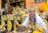 A woman in traditional attire sits in front of colorful spice pyramids, wearing a vibrant yellow dress with embroidered details. The scene is set in a market with containers of various spices arranged in the background.