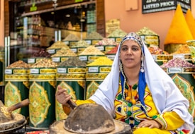 A woman in traditional attire sits in front of colorful spice pyramids, wearing a vibrant yellow dress with embroidered details. The scene is set in a market with containers of various spices arranged in the background.