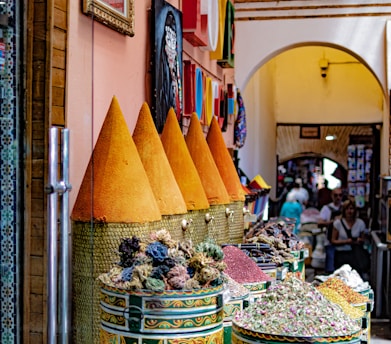 A vibrant market stall displays colorful cones of spices and dried flowers in decorative containers. The scene is bustling with activity, as people walk through a sunlit, arched passageway lined with paintings and textiles.