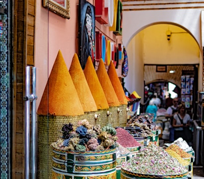 A vibrant market stall displays colorful cones of spices and dried flowers in decorative containers. The scene is bustling with activity, as people walk through a sunlit, arched passageway lined with paintings and textiles.