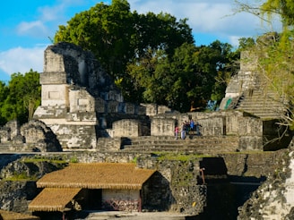 Ancient stone ruins are surrounded by lush green trees. The structures, including stepped platforms and thatched roofs, are built from weathered gray and white stone. A group of people is gathered on one of the platforms, suggesting a site of historical interest.