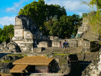 Ancient stone ruins are surrounded by lush green trees. The structures, including stepped platforms and thatched roofs, are built from weathered gray and white stone. A group of people is gathered on one of the platforms, suggesting a site of historical interest.