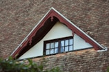 An architectural detail of a roof with a gable window. The roof is covered with reddish-brown, weathered shingles. The gable features decorative wooden trim painted in a muted red, framing a small window with white curtains. The window's dark frame contrasts with the light-colored wall. Green foliage is partially visible in the foreground.