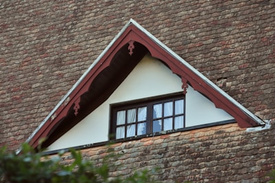 An architectural detail of a roof with a gable window. The roof is covered with reddish-brown, weathered shingles. The gable features decorative wooden trim painted in a muted red, framing a small window with white curtains. The window's dark frame contrasts with the light-colored wall. Green foliage is partially visible in the foreground.