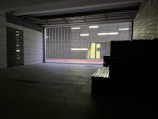 A dimly lit urban space with concrete walls and a metal security grille. Outside, there is a corridor with bright overhead lights and a vibrant yellow wall. A red strip runs along the base of the wall, adding a pop of color.