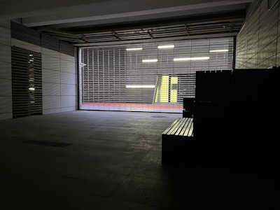 A dimly lit urban space with concrete walls and a metal security grille. Outside, there is a corridor with bright overhead lights and a vibrant yellow wall. A red strip runs along the base of the wall, adding a pop of color.