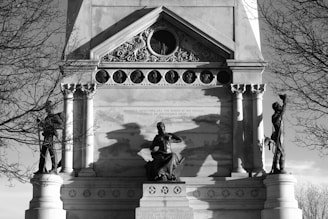 A historical monument featuring a sculptural group with three prominent statues. The central figure is seated with an authoritative posture, flanked by standing figures on each side. Ornate architectural details such as columns and decorative carvings frame the scene. The structure is set against a backdrop of bare trees, and the shadows add a dramatic element to the composition.