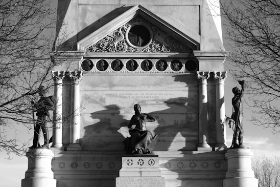 A historical monument featuring a sculptural group with three prominent statues. The central figure is seated with an authoritative posture, flanked by standing figures on each side. Ornate architectural details such as columns and decorative carvings frame the scene. The structure is set against a backdrop of bare trees, and the shadows add a dramatic element to the composition.