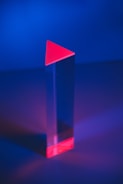 A tall, transparent prism with a triangular red top and base is set against a vibrant, deep blue background. The prism casts a subtle reflection on the surface below, creating a play of light and shadow.