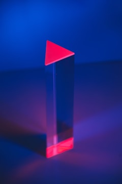 A tall, transparent prism with a triangular red top and base is set against a vibrant, deep blue background. The prism casts a subtle reflection on the surface below, creating a play of light and shadow.