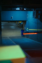A dimly lit parking garage with concrete floors and walls painted in blue. Fluorescent lights are visibly casting a cool glow on the surfaces. A few cars are parked in the distance, and there is a red barrier arm across an entrance or exit.