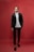 A person stands confidently against a solid red background, dressed in a stylish black suit with a white turtleneck. The outfit is completed with black loafers, and the individual has neatly groomed hair.