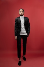 A person stands confidently against a solid red background, dressed in a stylish black suit with a white turtleneck. The outfit is completed with black loafers, and the individual has neatly groomed hair.