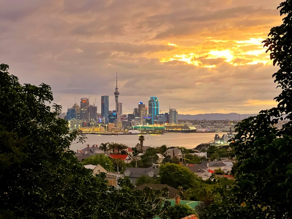 A vibrant city skyline at sunset, illuminated by the golden hues of the sky. Skyscrapers with glowing lights are prominent, with a mix of modern and traditional architecture. The foreground features lush green trees framing the city, with a body of water separating the urban landscape from the residential area in the foreground.