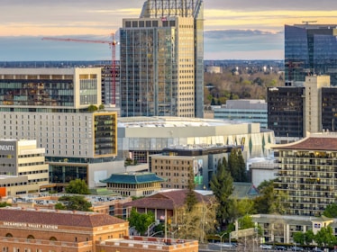 A cityscape featuring several modern high-rise buildings and an office tower with reflective glass windows. In the foreground, an older brick building labeled Sacramento Valley Station adds a historical element. A construction crane can be seen against the skyline, indicating ongoing development. The scene is set during a late afternoon, casting a warm glow on the structures.