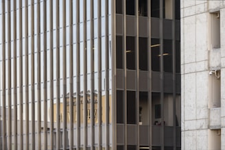 A grid of reflective glass windows dominates the scene, with vertical metal frames adding structure. The reflection of another building is visible, its features distorted and elongated due to the glass surface. Adjacent to the grid is a section of concrete wall, adding contrast with its solid texture and neutral color.