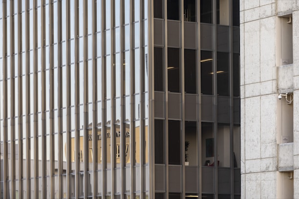A grid of reflective glass windows dominates the scene, with vertical metal frames adding structure. The reflection of another building is visible, its features distorted and elongated due to the glass surface. Adjacent to the grid is a section of concrete wall, adding contrast with its solid texture and neutral color.