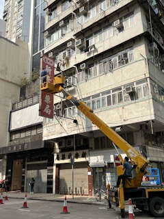 A worker operates a yellow cherry picker lift in front of a multi-story building. The building exterior features numerous air conditioning units attached to the windows, with some laundry hanging outside. The worker appears to be repairing or installing a signboard with Chinese characters. Below, traffic cones are placed on the street, and a few pedestrians walk by.