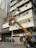 A worker operates a yellow cherry picker lift in front of a multi-story building. The building exterior features numerous air conditioning units attached to the windows, with some laundry hanging outside. The worker appears to be repairing or installing a signboard with Chinese characters. Below, traffic cones are placed on the street, and a few pedestrians walk by.