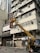 A worker operates a yellow cherry picker lift in front of a multi-story building. The building exterior features numerous air conditioning units attached to the windows, with some laundry hanging outside. The worker appears to be repairing or installing a signboard with Chinese characters. Below, traffic cones are placed on the street, and a few pedestrians walk by.