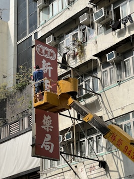A person stands on a raised platform from a yellow crane, adjusting a red sign with white Chinese characters on the side of an older apartment building. The building facade is weathered, with several open windows, air conditioning units, and clothes drying outside.