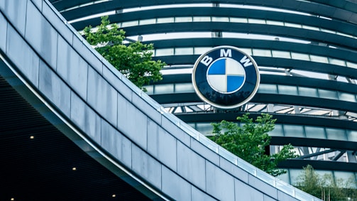 An architectural structure with curved, metallic elements is shown, prominently featuring the BMW logo against a backdrop of blue-tinted glass and gray steel beams. Green trees add a touch of nature to the sleek, modern design.