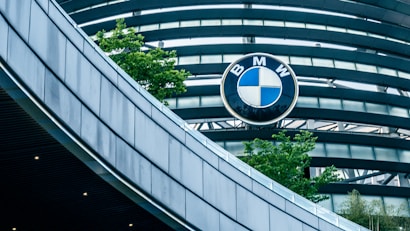 An architectural structure with curved, metallic elements is shown, prominently featuring the BMW logo against a backdrop of blue-tinted glass and gray steel beams. Green trees add a touch of nature to the sleek, modern design.