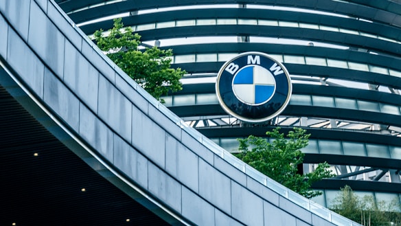 An architectural structure with curved, metallic elements is shown, prominently featuring the BMW logo against a backdrop of blue-tinted glass and gray steel beams. Green trees add a touch of nature to the sleek, modern design.
