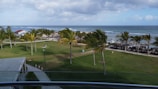 A coastal resort area with a view of the sea. The foreground includes a grassy area with palm trees scattered around. There is a covered walkway and a small building with a red roof to the left. In the background, there are beach umbrellas and chairs lined up along the shore. The sky is partly cloudy with a mix of blue and gray tones.