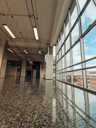A modern hallway with large floor-to-ceiling windows on the right side, allowing natural light to flood the space. The polished terrazzo floor reflects the light and the ceiling fixtures. Overhead, exposed pipes and fluorescent lights run along the ceiling, adding an industrial touch to the sleek and open architecture.