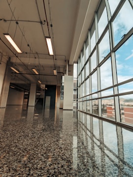 A modern hallway with large floor-to-ceiling windows on the right side, allowing natural light to flood the space. The polished terrazzo floor reflects the light and the ceiling fixtures. Overhead, exposed pipes and fluorescent lights run along the ceiling, adding an industrial touch to the sleek and open architecture.