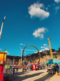 A vibrant amusement park scene with various rides and attractions under a clear blue sky with scattered fluffy clouds. Prominent features include a large, colorful circular ride and multiple smaller tents and booths. People are walking around, enjoying the lively atmosphere.