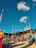 A vibrant amusement park scene with various rides and attractions under a clear blue sky with scattered fluffy clouds. Prominent features include a large, colorful circular ride and multiple smaller tents and booths. People are walking around, enjoying the lively atmosphere.