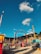 A vibrant amusement park scene with various rides and attractions under a clear blue sky with scattered fluffy clouds. Prominent features include a large, colorful circular ride and multiple smaller tents and booths. People are walking around, enjoying the lively atmosphere.