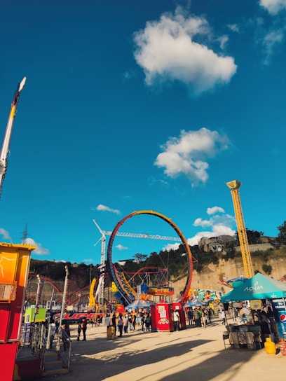 A vibrant amusement park scene with various rides and attractions under a clear blue sky with scattered fluffy clouds. Prominent features include a large, colorful circular ride and multiple smaller tents and booths. People are walking around, enjoying the lively atmosphere.