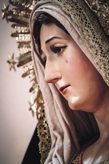 A close-up view of a statue depicting a woman's face, adorned with a delicate headscarf and golden decorations. The woman has a tear rolling down her cheek, and her expression appears somber and reflective.