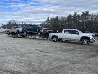 A large silver pickup truck is towing a trailer with two other pickup trucks secured on it. The setting appears to be a parking lot with a row of trees in the background and a cloudy sky above.