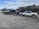 A large silver pickup truck is towing a trailer with two other pickup trucks secured on it. The setting appears to be a parking lot with a row of trees in the background and a cloudy sky above.