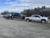 A large silver pickup truck is towing a trailer with two other pickup trucks secured on it. The setting appears to be a parking lot with a row of trees in the background and a cloudy sky above.