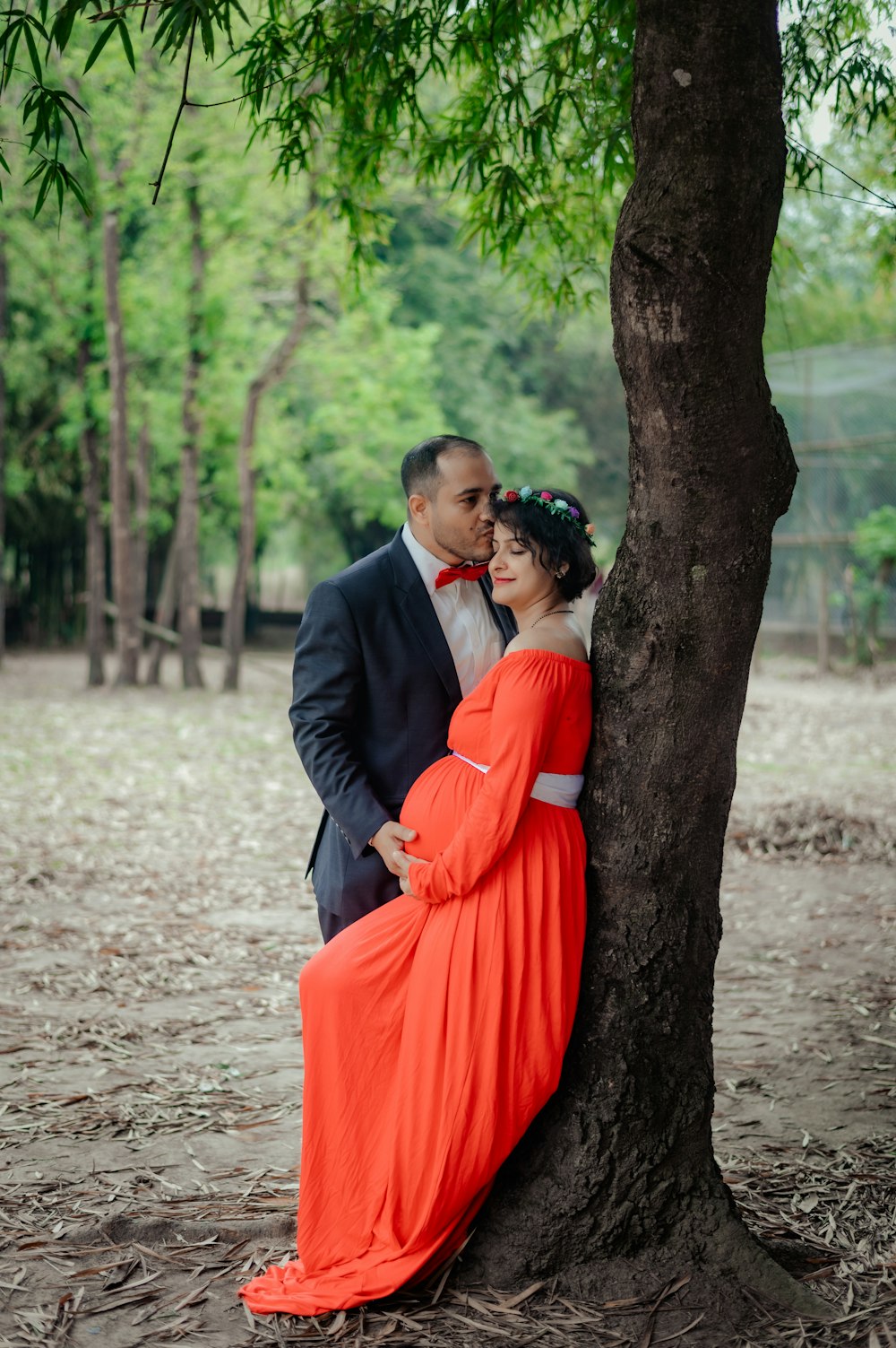 a man in a suit and woman in an orange dress leaning against a tree