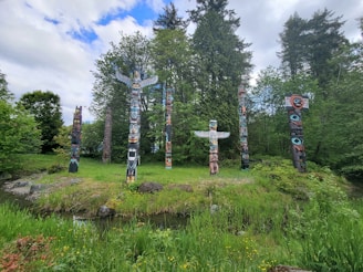 A wide-angle photo capturing a diverse group of totem poles in a natural forest setting, surrounded by lush greenery.
