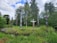 A group of intricately carved totem poles stand in a lush, green forested area. The totem poles display colorful and detailed designs, featuring various symbols and figures. The surrounding landscape includes vibrant grass, tall trees, and a partially cloudy sky.