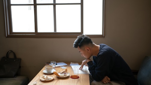 A person is seated at a wooden table near a window, focused on reading a document. The table is cluttered with a plate of pastries, a cup of coffee, a glass of water, and some papers. The setting appears calm and studious, with soft lighting coming through the window.