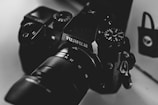 A close-up, monochrome photograph of a Fujifilm camera with visible dials and lens details. The image exhibits a smooth texture and high contrast, highlighting the camera's intricate design and branding.