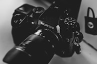A close-up, monochrome photograph of a Fujifilm camera with visible dials and lens details. The image exhibits a smooth texture and high contrast, highlighting the camera's intricate design and branding.