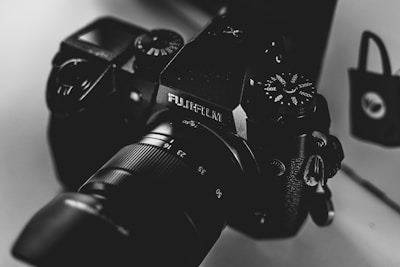 A close-up, monochrome photograph of a Fujifilm camera with visible dials and lens details. The image exhibits a smooth texture and high contrast, highlighting the camera's intricate design and branding.
