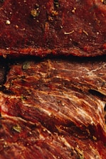 Several pieces of dried and seasoned meat with a reddish-brown hue are shown. The meat features visible textures and fibers, along with various herbs and spices sprinkled across the surface.