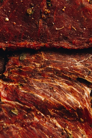 Several pieces of dried and seasoned meat with a reddish-brown hue are shown. The meat features visible textures and fibers, along with various herbs and spices sprinkled across the surface.