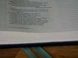 An open book displaying text with a focus on certain passages that discuss spiritual and moral themes. The page is light-colored with black printed text and a teal ribbon bookmark partially visible at the bottom. The book rests on a wooden surface.